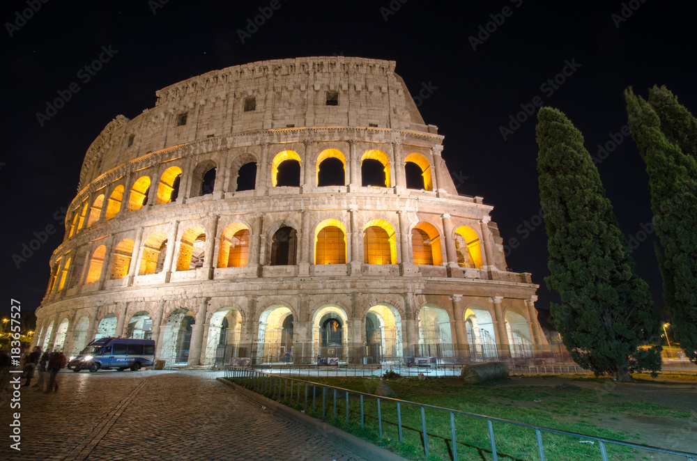A night image of the Colosseum with stars above. A police van sits out front with a crowd of tourists in the forground.