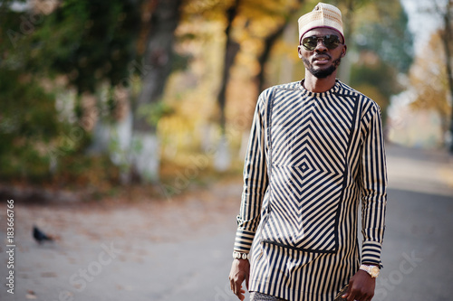 Portrait of stylish black african american man at hat and sunglasses against sunny autumn fall background. Rich people in africa at traditional dress.