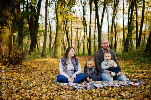 Happy caucasian family of mom dad and little girl with boy sitting on plaid at majestic autumn fall forest.