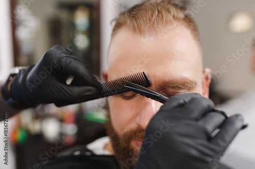 Male professional hairdresser serving client, shearing eyebrows with scissors. Ginger stylish young man with thick big beard and short hair getting trendy haircut in black cape. Light barber shop room © ViDi Studio