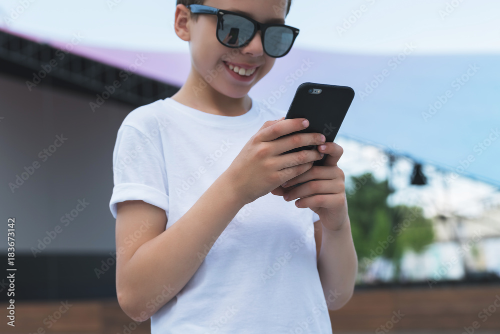 Summer day. Smiling boy in white T-shirt and sunglasses stands outdoor and uses smartphone. Boy plays computer games on digital gadget, surfing internet. Social networks, computer games. Summer day. Smiling boy in white T-shirt and sunglasses stands outdoor and uses smartphone. Boy plays computer games on digital gadget, surfing internet. Social networks, computer games.