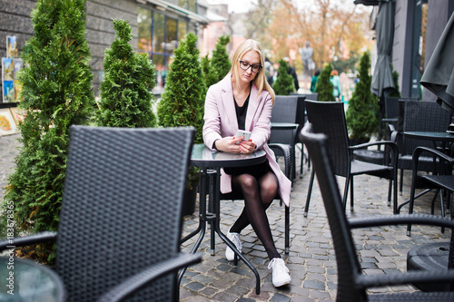 Blonde girl at glasses and pink coat, black tunic sitting at table outdoor cafe and speaking on phone.