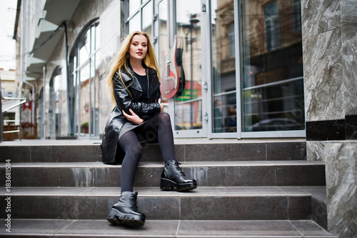 Blonde fashionable girl in long black leather coat posed at stairs.