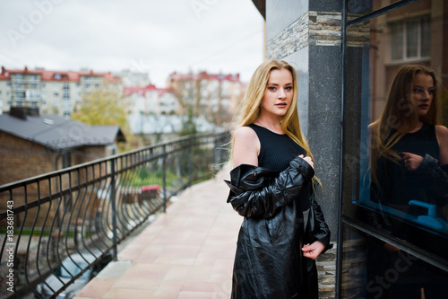 Blonde fashionable girl in long black leather coat posed against large window of building.
