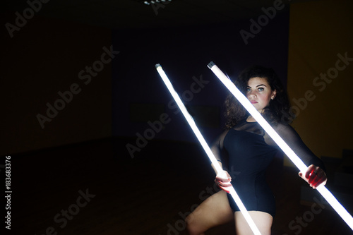 Curly brunette plus size model with two long led lamp at black room.