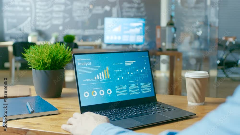 Male Analyst at His Desk Works on a Laptop Showing Statistics, Graphs and Charts. He Works in Light and Modern Creative Office.RED EPIC-W 8K Camera.