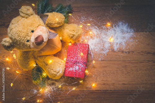 Cute teddy bear wrapped in shining golden Christmas ligts with a red gift box and pine tree branches on wooden background. Christmas joy or giving concept photo