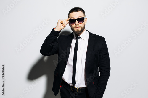 Portrait of a concentrated handsome man dressed in suit and hands on sunglasses looking away while standing over gray background