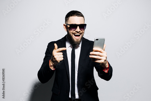 Portrait of a cheerful happy man in suit and tie read sms on phone and smile isolated over gray background