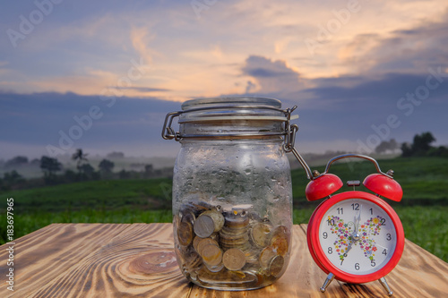 Obraz na płótnie Coins in jar  and alar clock on the pine wood  with beautiful sunrise as background for travel concept