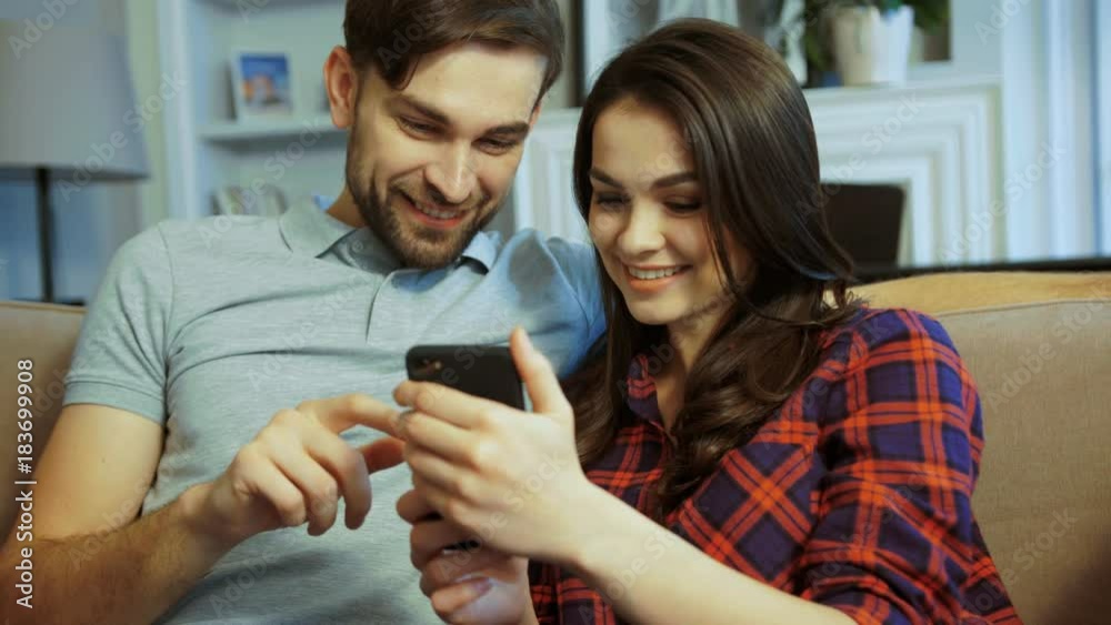 Portrait of young couple sitting on the sofa and scrolling on the smart phone in the cozy living room at home. Indoors. Close up Portrait of young couple sitting on the sofa and scrolling on the smart phone in the cozy living room at home. Indoors. Close up