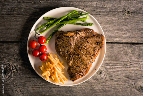 Cooked and plated steak on wood background with fries, tomato and asparagus