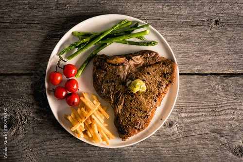 Cooked and plated steak on wood background with fries, tomato, asparagus and sage butter