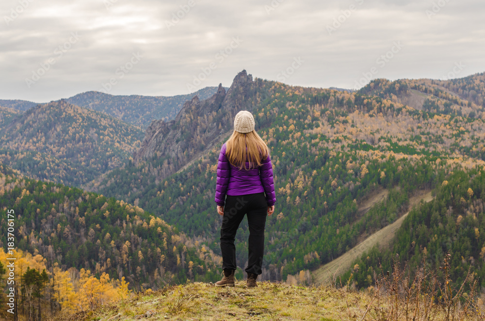 A girl in a lilac jacket standing on a mountain, a view of the mountains and an autumn forest by a cloudy day
