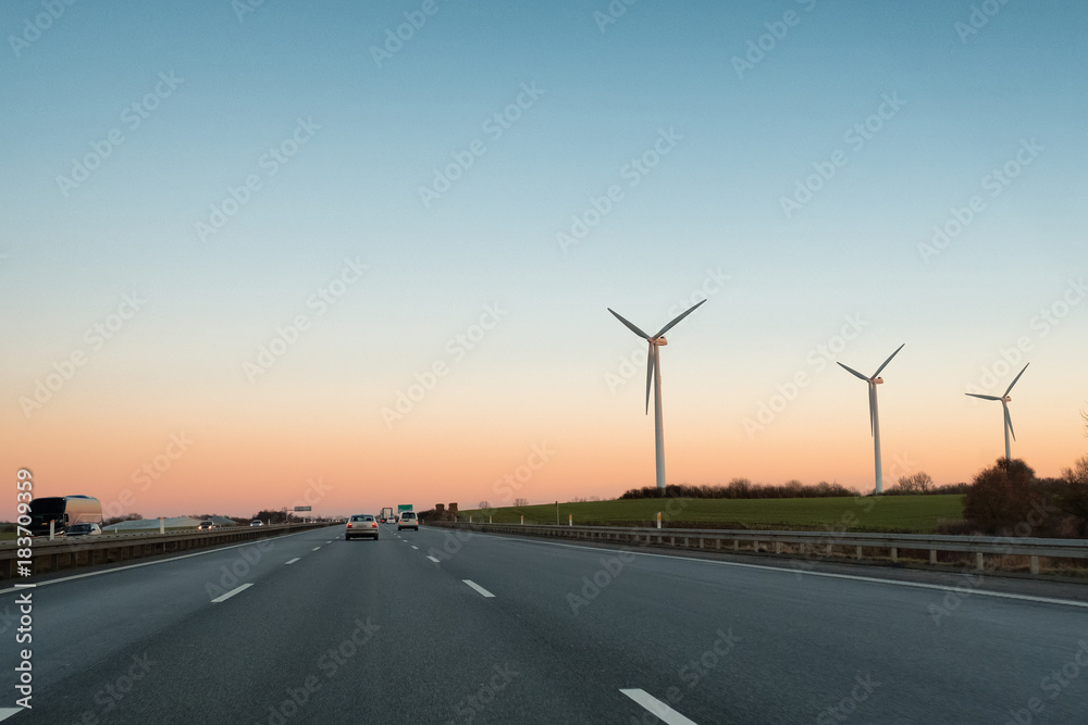 Multi-lane expressway at sunset. Highway. Three wind power plant generators are to the right of the road. Multi-lane expressway at sunset. Highway. Three wind power plant generators are to the right of the road.