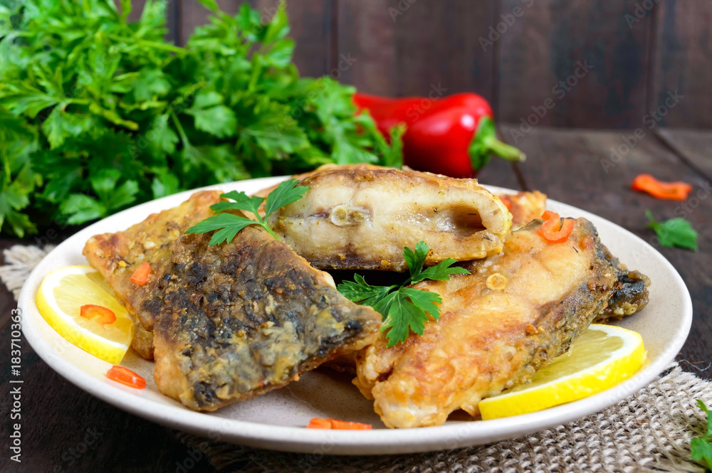 Pieces of roasted carp with lemon and greens on a plate on a dark wooden background. Pieces of roasted carp with lemon and greens on a plate on a dark wooden background.