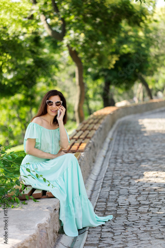 Young beautiful woman in a light green pastel long dress is sitting on a park bench and talking on a mobile phone.