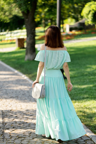 Young beautiful woman in a light green pastel long dress is walking in the city park in summer.