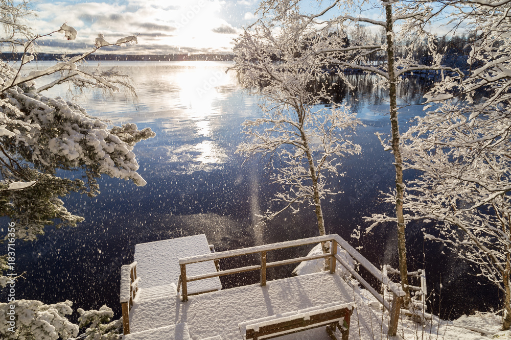 Beautiful view of Lake Pyhäjärvi, snowy trees and pier and snow falling from trees on a sunny day in the winter in Tampere, Finland.