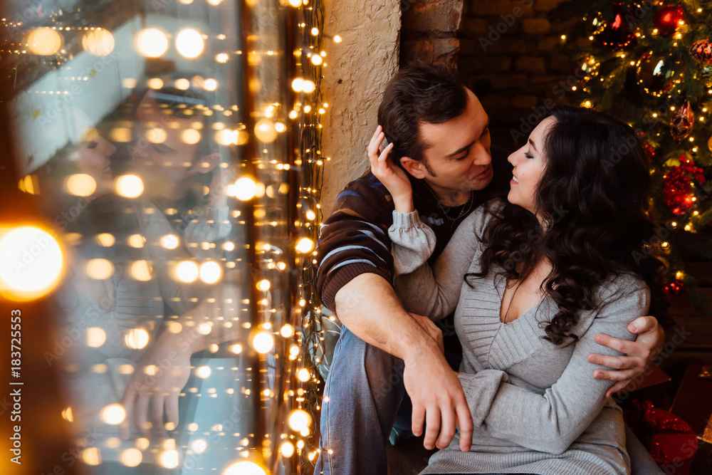 Couple of lovers hugs sitting on the windowsill in the Christmas loft studio. guy is hugging the girl. Couple of lovers hugs sitting on the windowsill in the Christmas loft studio. guy is hugging the girl.