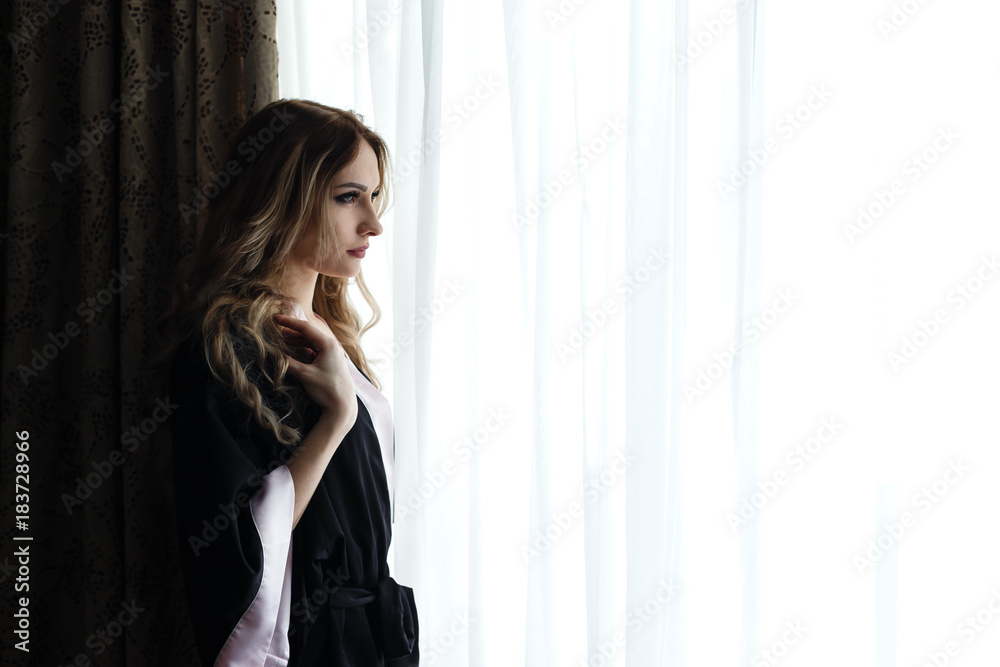 Beautiful young woman dressed in robe standing near a window in hotel. Beautiful young woman dressed in robe standing near a window in hotel.