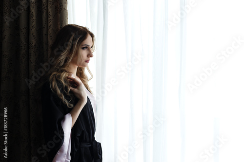 Beautiful young woman dressed in robe standing near a window in hotel.