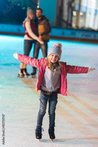 adorable little girl standing with open arms and smiling at camera while parents standing behind on rink