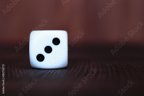 Single white plastic dice on brown wooden board background. Six side cube with black dots. Number 3.