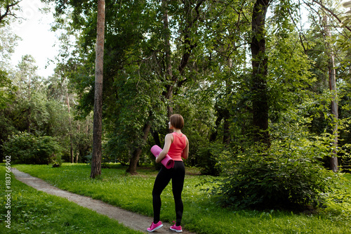 Training in the fresh air in the park. The sports girl in a pink undershirt against the background of green foliage. The woman holds in hand a rug for yoga. Portrait to the utmost from a back.