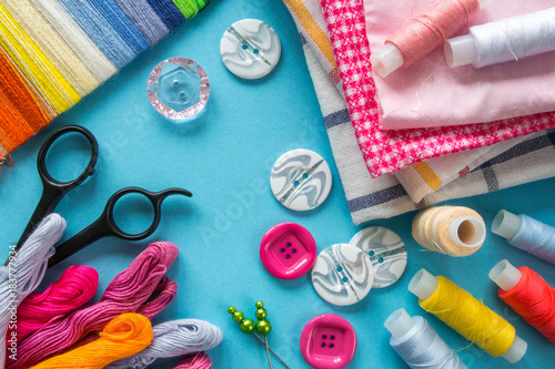 Multicolored threads, scissors, buttons, fabric and various sewing accessories on a blue background with copy space flat lay