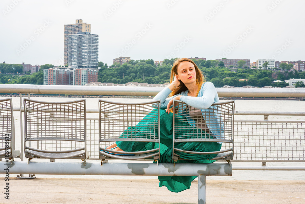 Lonely girl missing you. Wearing light blue sweater, green skirt, an American woman casually sitting on metal chair by Hudson River in New York, opposite New Jersey, looking tired, lost in thought..