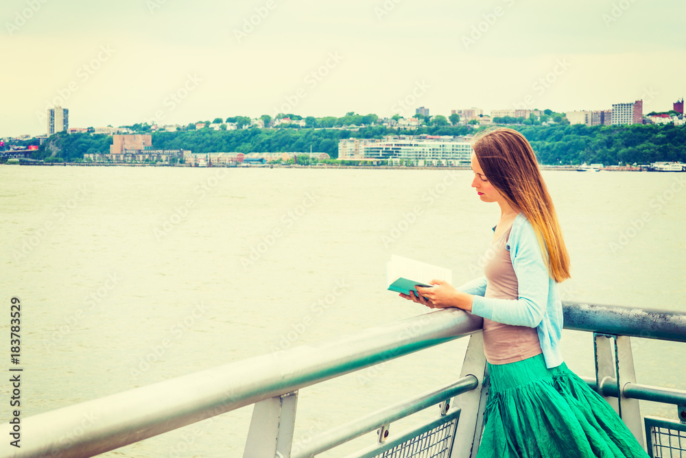 American college student studying, traveling in New York. A pretty girl wearing light blue cardigan, green skirt, standing by Hudson River, looking down, reading book. ..