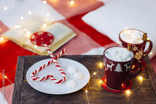 cups of cocoa with marshmallow on the wood box, hart candy cane on white plate, book on red carpet, garland, new year and chirstmas evening. homely chiiling evening time love to celebration holyday photo