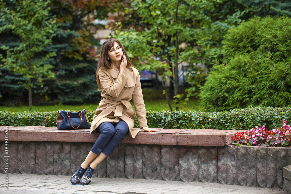 Portrait of happy young brunette woman in beige coat talking on the phone