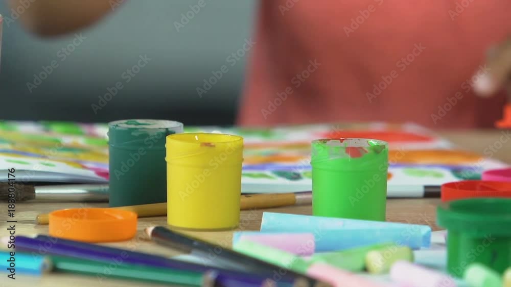 Table filled with acrylic paint and brushes, children drawing in background Table filled with acrylic paint and brushes, children drawing in background