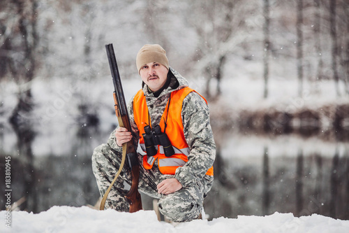 Male hunter in camouflage, armed with a rifle, sittiing in a snowy winter forest