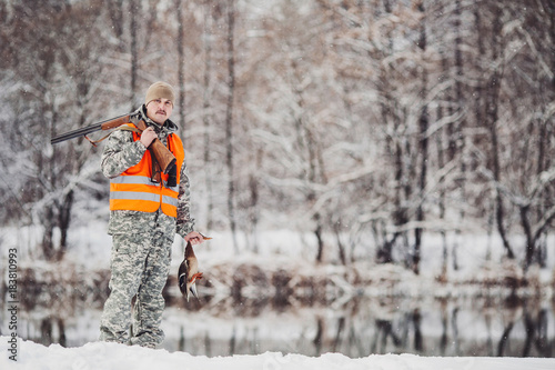 Male hunter in camouflage, armed with a rifle, standing in a snowy winter forest with duck prey