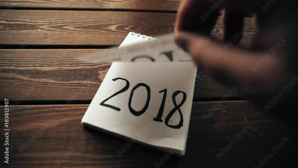 New Year 2018 is coming concept. Hand flips notepad sheet on wooden table. 2017 is turning, 2018 is opening
