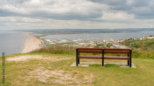 A bench on the South West Coast Path with view towards Fortuneswell and Chesil Beach, Isle of Portland, Jurassic Coast, Dorset, UK - with clouds over Weymouth in the background
