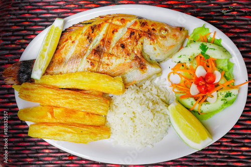 Above view of fried fish served with side salad, rice, and yucca served in a white plate over a wooden table photo