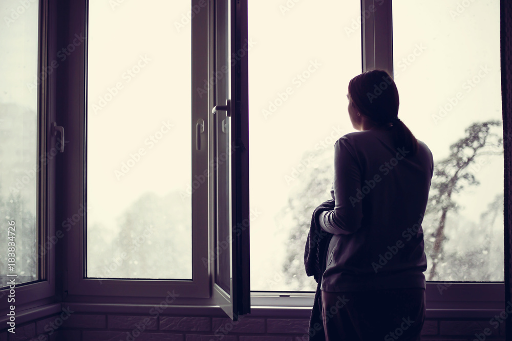 The girl is standing at the window and looks at how it's snowing