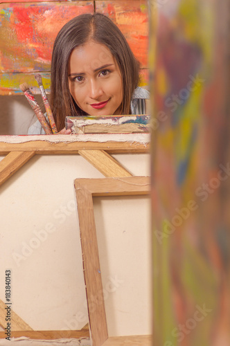 Beautiful woman painter holding paint brush and hiding in empty frame  in a studio paint background  art classes for adults  education concept