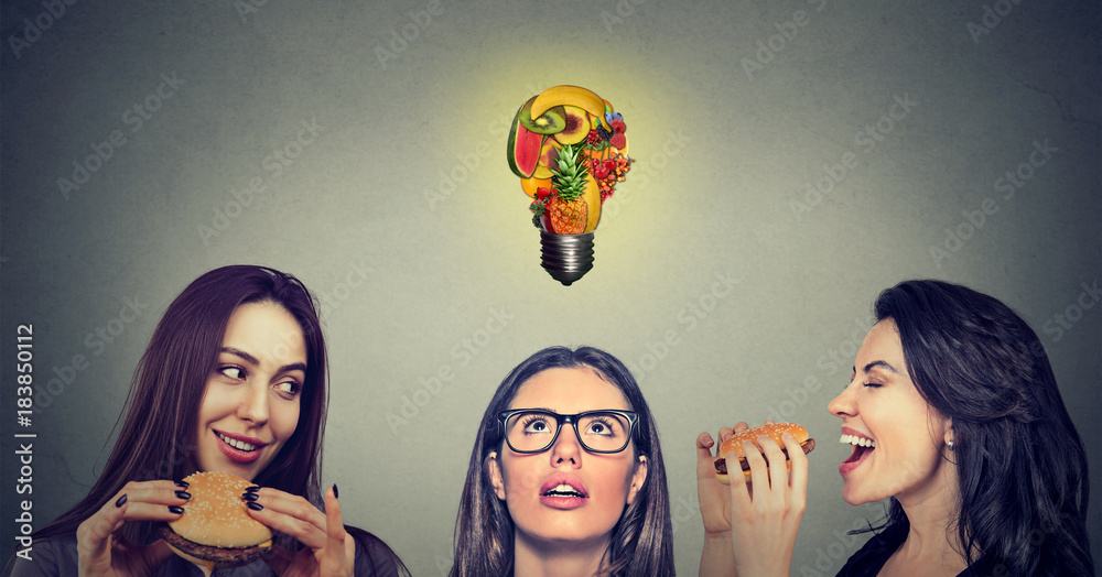 Φωτογραφία Two women eating hamburgers looking at stressed thoughtful girl with fruit shaped light bulb above head