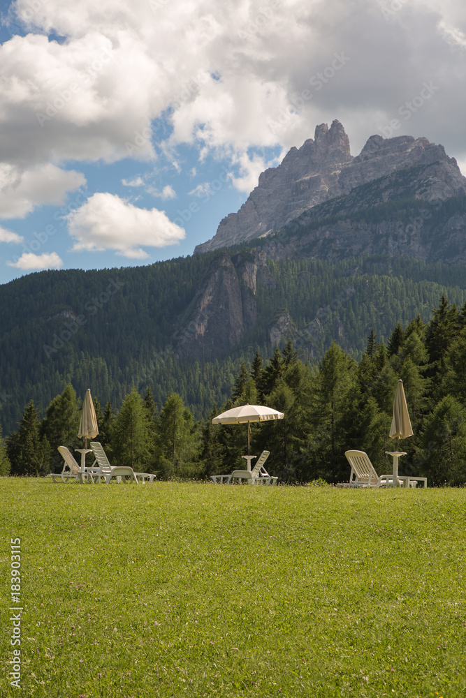 White Sun Loungers and Sun Umbrellas on Green Meadow in Italian Dolomites Alps Scenery