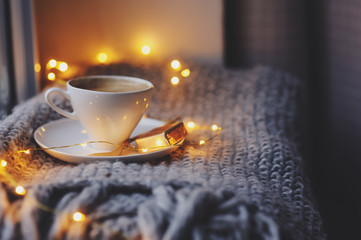 cozy winter or autumn morning at home. Hot coffee with gold metallic spoon, warm blanket, garland and candle lights, swedish hygge concept.