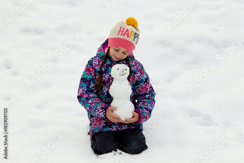 Happy little girl sitting on a snow in winter outdoors and tenderly holding funny snowman in her hands photo