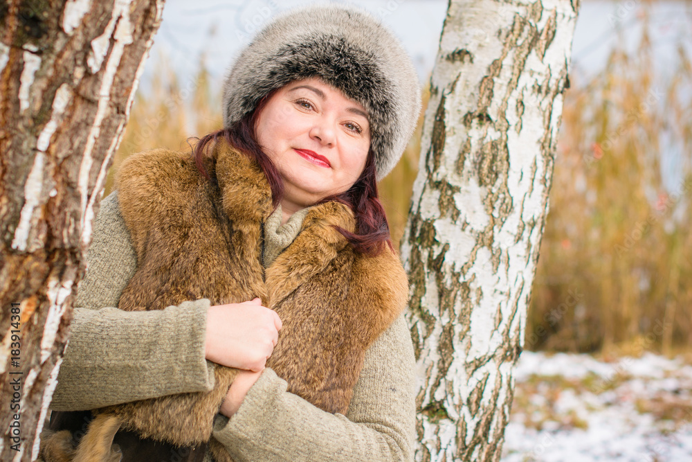 Plus size lady in fur coat wearing hat with snow and nature of Canada in the background. Portrait of smiling plump american woman in grey fur hat. Plus size lady in fur coat wearing hat with snow and nature of Canada in the background. Portrait of smiling plump american woman in grey fur hat.