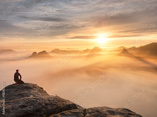 Tourist sit on peak of sandstone rock and watching into colorful mist and fog in  morning valley. Sad man