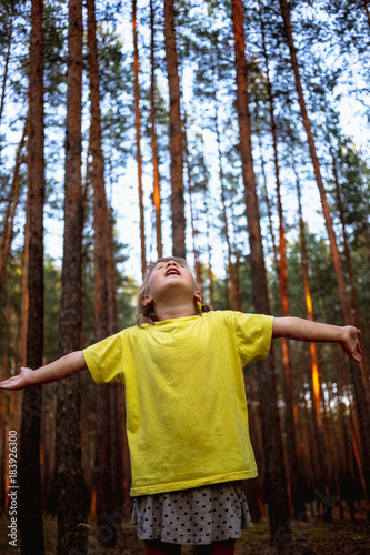 Beautiful little girl in pine forest with hands up enjoys nature.