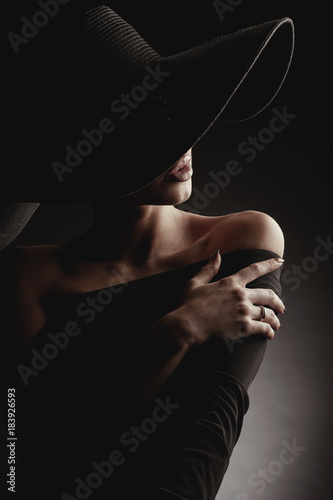Dark studio portrait of elegant woman in black wide hat and black dress. Hidden half face.
